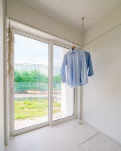 笛吹市の新築実例。大きなテラス窓と高窓から光が差し込む、室内干し専用のサンルーム。白で統一された空間に、洗濯物を干したシャツが掛かる清潔感のあるランドリールーム。